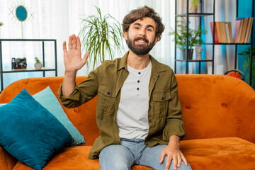 Fototapeta premium Hello. Middle eastern handsome man smiling friendly at camera and waving hands gesturing hello hi, greeting or goodbye, welcoming with hospitable expression at home. Guy sitting on sofa in living room