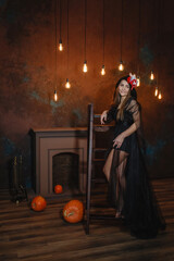 A beautiful woman in a black witch costume stands near the stairs by the fireplace.