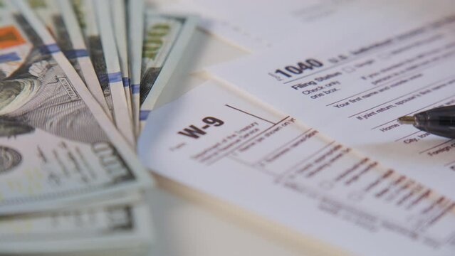 American dollar tax bill, pen calculator and U.S. money. 1040 and W9 individual tax form are placed on desk of American citizen. rotate camera shot
