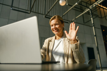 Woman freelancer talking with client via video call and waving Hi while sitting in office - obrazy, fototapety, plakaty