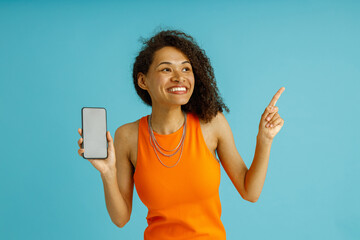 Positive woman with mobile phone points at side, looks amazed by promo offer on blue background