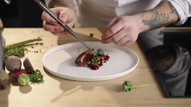 Restaurant chef putting fresh basil leaf on duck steak with sliced beetroot on white plate in slow-motion