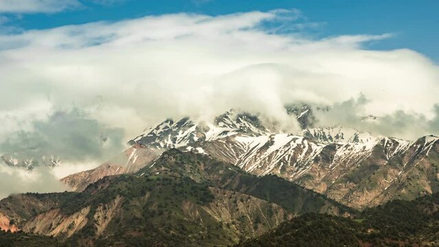 Timelapse mountains. Mountain range is covered with seething cap of white fluffy clouds. Clouds cling to snow-capped mountains peaks. Summer blue sky time lapse. Cinematic background video