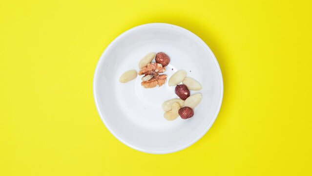 Nuts filling a bowl on a bright yellow background. Stop motion video with the cashews, hazelnuts, almonds nuts disappear from the white ceramic bowl.