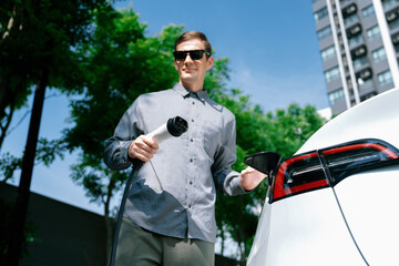 Young man travel with EV electric car charging in green sustainable city outdoor garden in summer...
