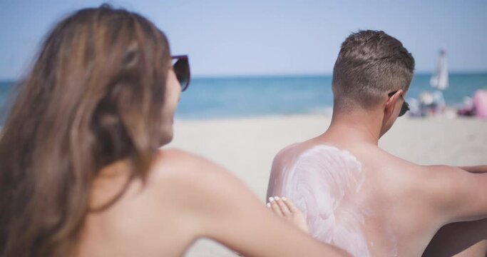 Couple on a sand beach applying suncream. Girl drawing heath on boyfriends back. Enjoying summer at sea concept.