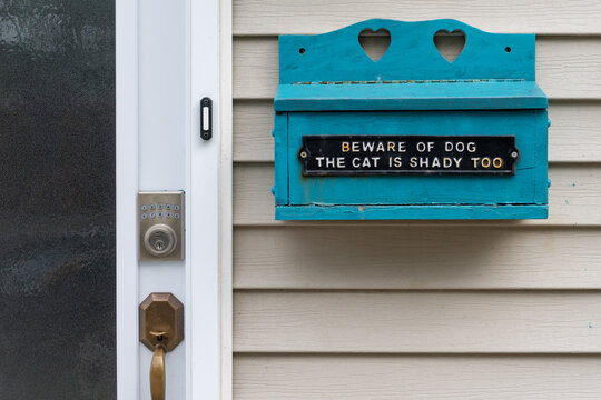 A vibrant blue wooden mailbox on the wall of a house, next to the entrance of a beige-colored house. The wood postal box has a black metal sign which says beware of the dog and the cat is shady too. 