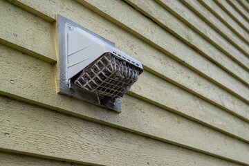 The exterior of a bright yellow house wall with a white dryer vent exposes the air vent which is clogged with hair, wood, and dirt in the mesh causing a fire hazard. The plastic unit guard is dirty.