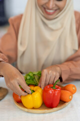 A young Muslim woman in a hijab prepares colorful vegetables on a wooden plate. Useful for making healthy food menus, vegetable salads, while preparing for online blogger broadcasts in the kitchen.
