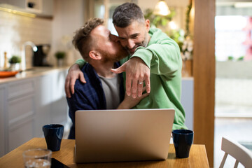 Young male gay couple celebrating getting engaged and showing off the ring over a video call on the laptop in the living room