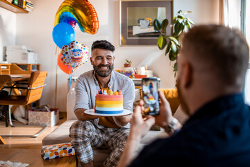 Young gay male couple celebrating a birthday at home and taking a photo with a phone