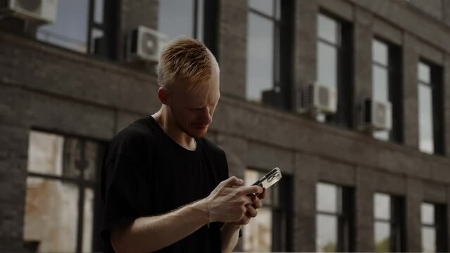 A man chooses music on a smartphone. A young man in a black T-shirt stands in front of a building. Young man in black t-shirt with mobile phone in the city