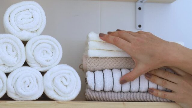 Women's hands place a pile of clean towels in neutral and white colors on a wooden shelf.