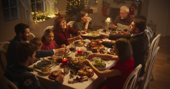 Diverse Group of People, Young and Old, Share a Joyful Christmas Dinner at Home. Family Sitting Behind a Big Dining Table Full of Delicious Food with Roast Turkey Feast. High Angle Evening Footage