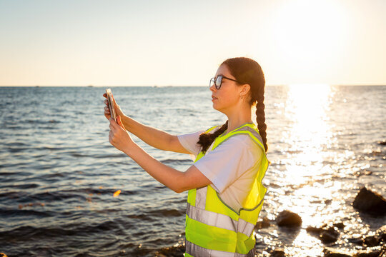 Inspection environmentalist. Side view of young Caucasian woman wearing glasses using tablet analyzing water in ocean. In background is ocean, coast and sunset. Concept of environmental protection