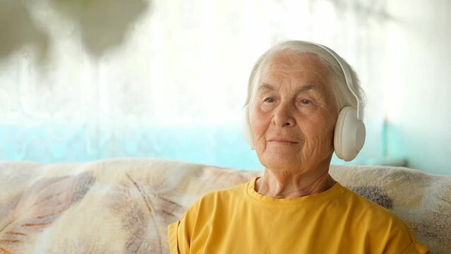 Close-up of an elderly woman with gray hair wearing headphones sitting at home on the couch and listening to music or an audiobook