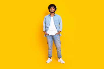 Fototapeta na wymiar Full size photo of satisfied cool man with curly hairdo dressed denim shirt jeans hold arms in pockets isolated on yellow color background