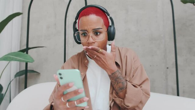 Young shocked African American woman fearfully covers mouth with hand after reading amazing news on mobile phone or receiving SMS message about bank bankruptcy sits on sofa in home. Problems, troubles