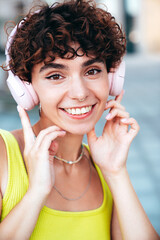 Young beautiful smiling hipster woman in trendy summer clothes. Carefree woman with curls hairstyle, posing in the street at sunny day. Positive model outdoors. Listens music at her earphones