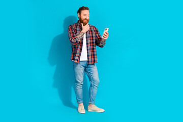 Full size photo of minded thoughtful man with tattoo wear plaid shirt denim trousers look at smartphone isolated on blue color background