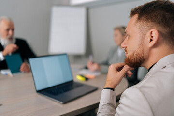 View from shoulder of thoughtful businessman sit at desk with colleagues in office talk discuss business ideas at briefing together. Pensive businesspeople brainstorm cooperate using laptop at meeting