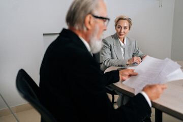 Smiling middle-aged CEO businesswoman in suit looking listening to senior adult executive manager discussing corporation financial plan at office table holding documents, teamwork corporate meeting.