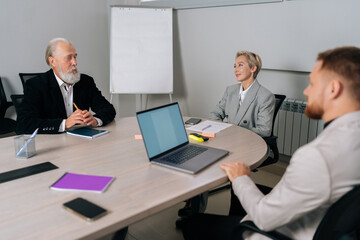 Positive middle-aged businesswoman attending meeting with diverse-ages colleagues in office. Experienced business woman smiling sitting with team in meeting room, working together.