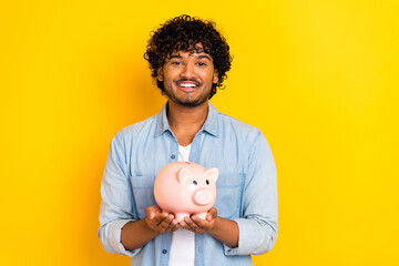 Portrait of cheerful wealthy person toothy smile arms hold money pig bank isolated on yellow color...