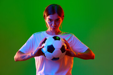 Motivated and competitive young girl, football player posing with ball against green studio...