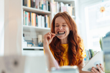 Happy woman biting credit card and holding tablet PC at home