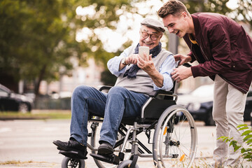 Laughing senior man in wheelchair and his adult grandson looking together at smartphone having fun