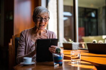 Mature woman is sitting in cafe and relaxing. She is using digital tablet.