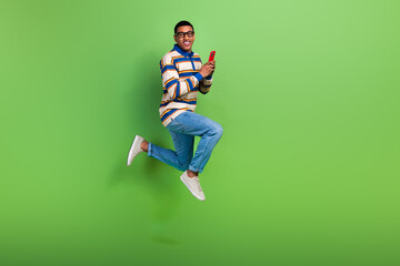 Fototapeta na wymiar Full size body photo of crazy activity jumping guy influencer with smartphone wear jeans browsing phone isolated on green color background