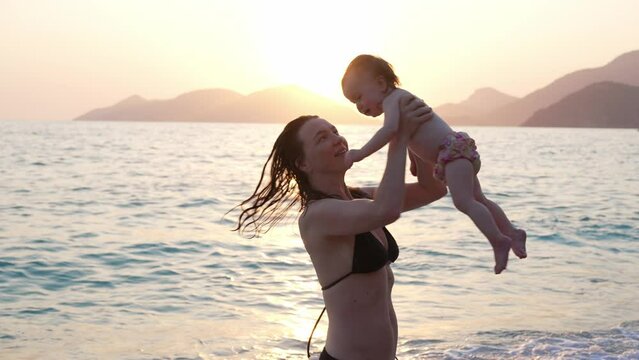 Young Mother in Swim Suit Playing and Hugging her Newborn Baby on Sea Beach