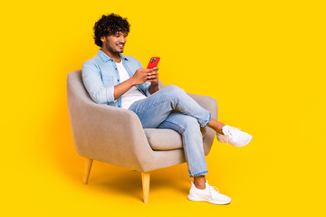 Full length photo of smart clever man wear jeans shirt in armchair hold smartphone read notification isolated on yellow color background