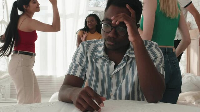 Sad upset depressed african black male having headache during fun dance party, friends dancing on background. Cultural shock, misunderstanding, language barrier. Apathetic multiethnic friends 