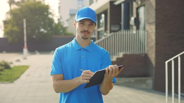 Potrait of Caucasian Male Real Estate Agent Looking at Camera Standing near Cottage City. Realtor Worker in Blue Uniform Holding Documents at a Tablet