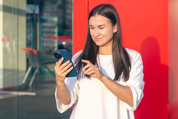 A young woman uses a smartphone, stands near a shopping center at sunset.