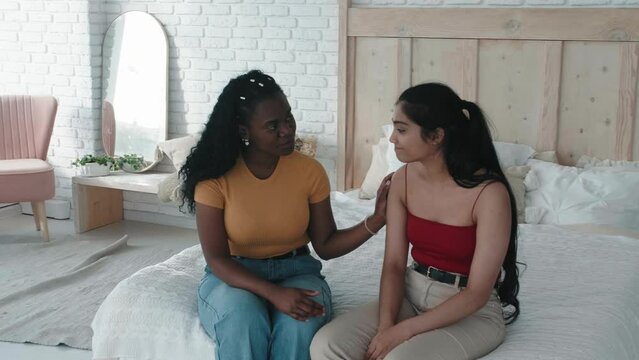 Female friendship. African American female talking to latin American girl, giving support and encouragement. Upset indian woman hugging her friend, sitting on bed at home. Loving caring black woman 