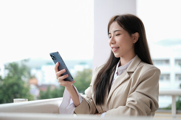 Satisfied asian businesswoman using mobile phone on office terrace with city view on background