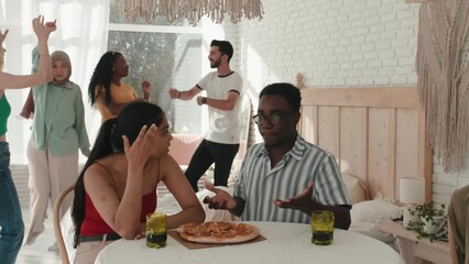 Funny multiethnic friends at party, dancing and eating pizza, african American man with pretty latin American chatting, eating pizza and cheering, clinking. Students have fun in campus. Diverse people