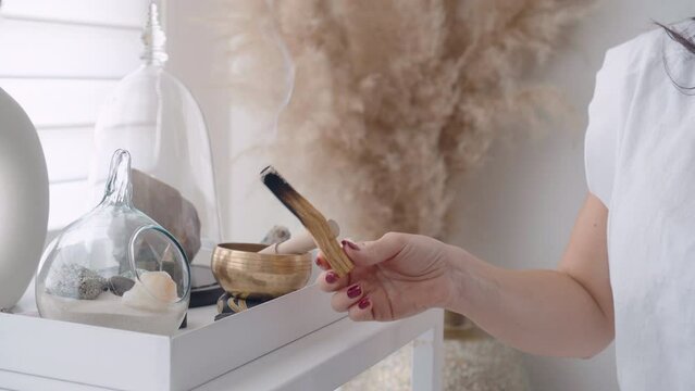 Woman holding Palo Santo stick and shaking it in the environment spreading the smoke and placing it on an abalone shell on an altar with quartz crystals preparing for the spiritual class of yoga