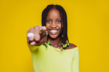 Young african woman points finger directly at camera with positive expression, says I choose you, wears casual outfit and braid hair, stands over yellow wall with blank space area. You are my type.