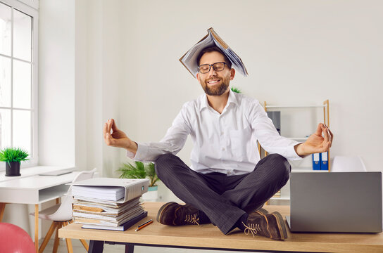 Portrait of a funny relaxed tired business man with a folder on head meditating and having a rest in the office or at home sitting on the table on his workplace in Lotus pose with closed eyes.