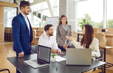 Team of four business people having corporate staff work meeting. Group of friendly young men and women using modern laptops and having discussion at wooden table in cozy office interior