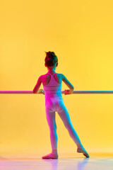Rear view portrait of small adorable ballerina dancer girl in ballet suit, legging and pointe practicing for performance in dance school in neon light