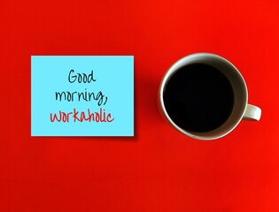 Coffee cup on orange background with blue note written GOOD MORNING WORKAHOLIC, concept of person who works excessively and compulsively, work too hard and is unable to detach from work