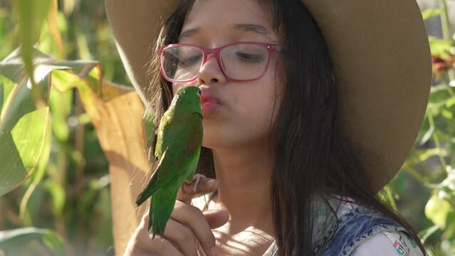 Little girl playing and kissing a green parakeet. Brotogeris jugularis. in the middle of a corn plantation, wearing a hat, coverall and glasses. Exotic animals.