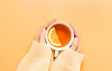 Hands in yellow sweater hold cup of tea with lemon, orange background, top view. Woman with  cup of tea, cozy autumn and winter concept, healthy lifestyle