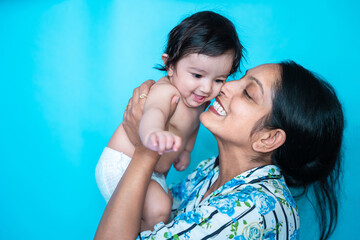 Smiling indian mother playing with her six months cute little baby in diaper isolated over blue background. Happy family. Asian mom with infant child having fun, Motherhood. Copy space. - obrazy, fototapety, plakaty